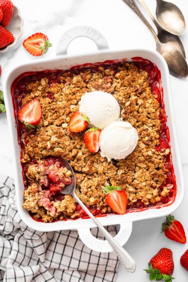 Baked strawberry crisp topped with two scoops of vanilla ice cream and fresh strawberry halves. There is a spoon scooping out some of the crisp.