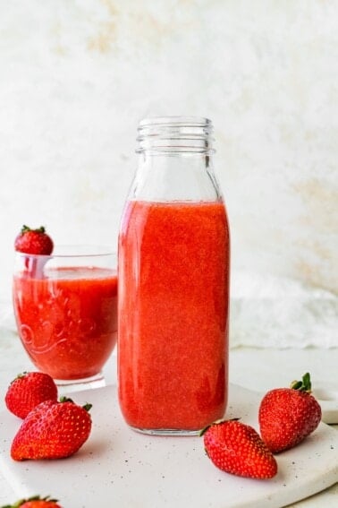 Glass bottle filled with strawberry juice placed on a white surface, with a glass of juice and strawberries beside it.