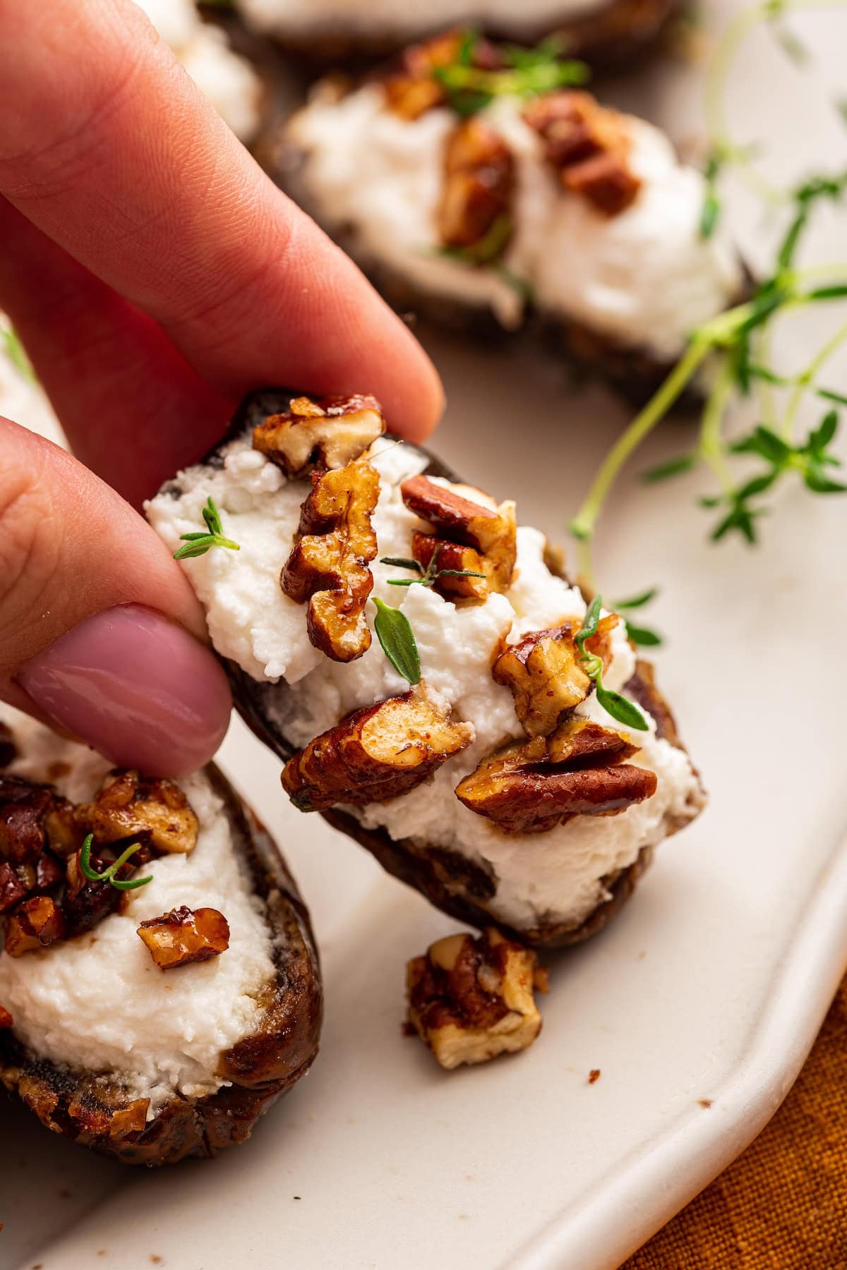 A womans hand is grabbing a date that is stuffed with goat cheese and topped with chopped pecans and fresh thyme.