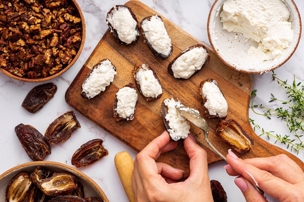 A womans hands uses a spoon to stuff halved dates with goat cheese.