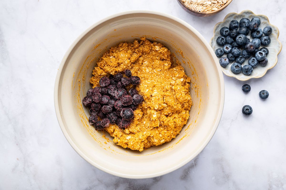 Frozen blueberries being added to the sweet potato blueberry oatmeal cookie dough in a mixing bowl.