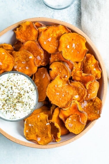 Baked homemade sweet potato chips on a bowl and topped with flakey salt. A bowl of herby creamy dip is beside the chips.