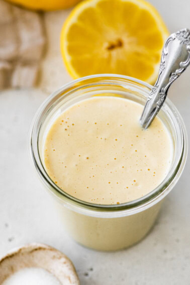 A creamy tahini dressing in a glass jar with a metal spoon and a halved lemon in the background.