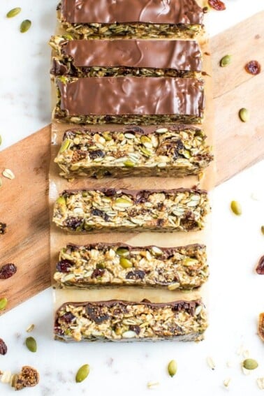 Slices of tahini fig bars topped with chocolate, slices on a cutting board and surrounded by pumpkin seeds and dried figs.
