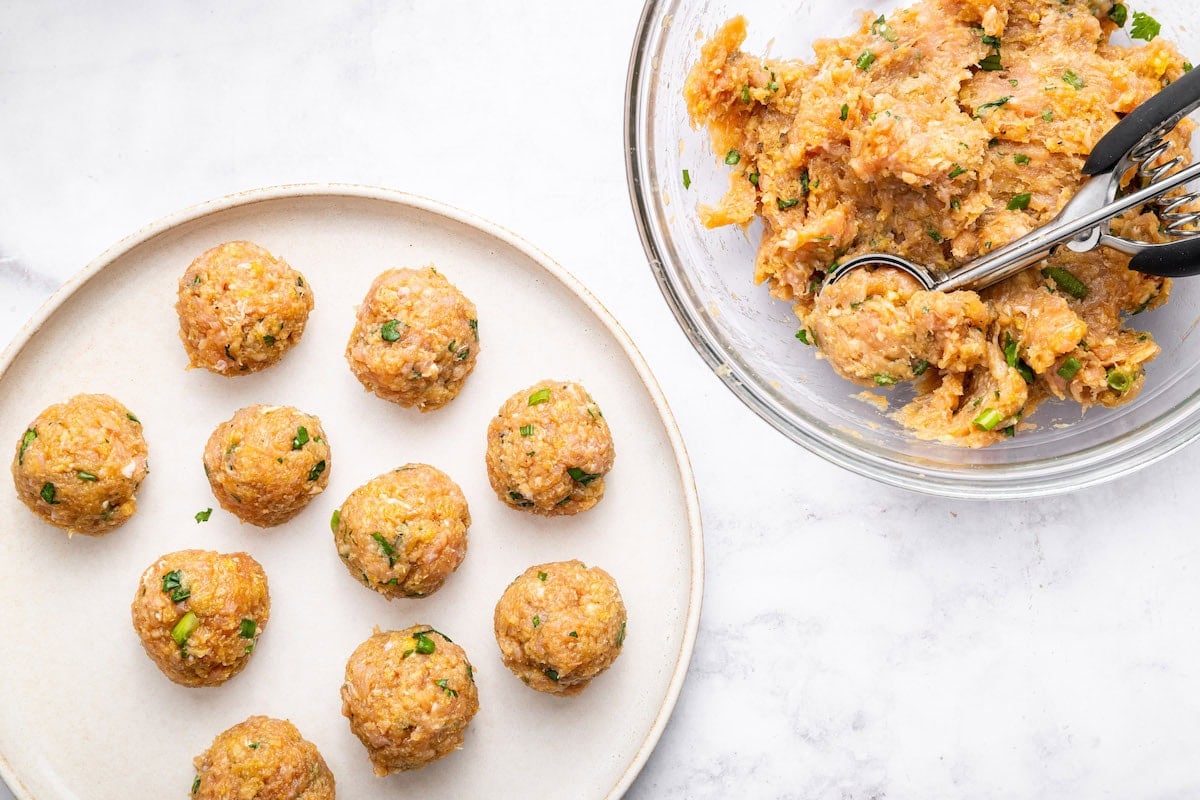 Thai chicken meatballs being formed using a cookie scoop. 10 meatballs are formed on a plate and the rest of the mixture is in a glass mixing bowl with a cookie scoop in it.