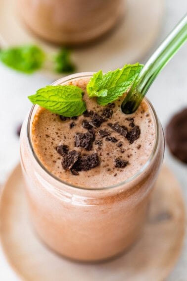 One mint chocolate protein shake in a glass topped with crushed Thin Mint cookies and fresh mint. There is a green straw in the glass.