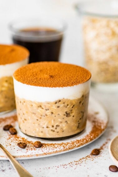 Glass jar of tiramisu overnight oats topped with Greek yogurt and cocoa powder, styled on a plate with coffee beans and spoons.