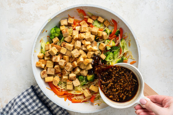 Tofu, cabbage, carrots, broccoli and red bell pepper in a skillet with woman's hand pouring sauce over ingredients.