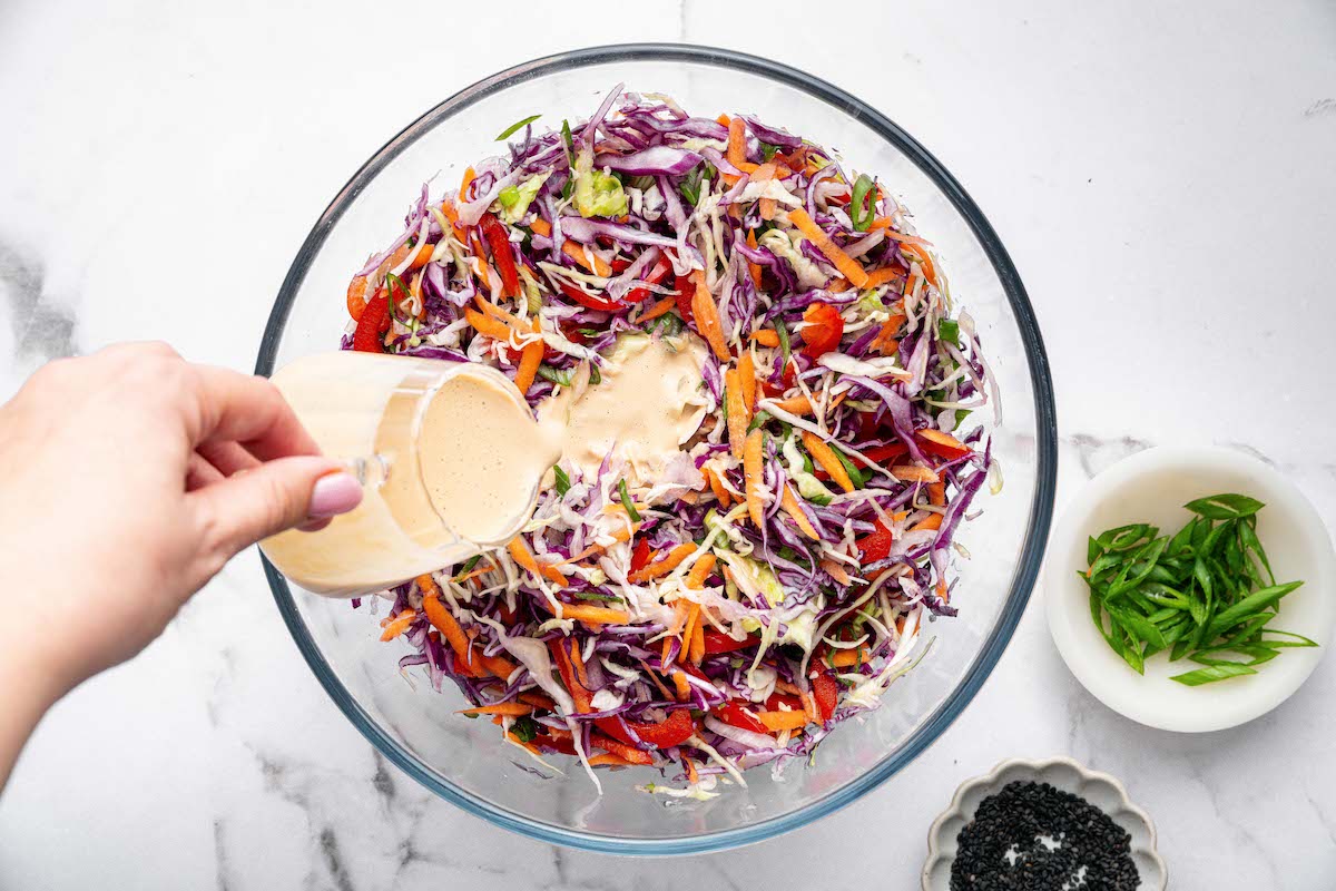 Pouring creamy sesame-ginger dressing over shredded cabbage, carrots, and bell pepper in a large glass mixing bowl.