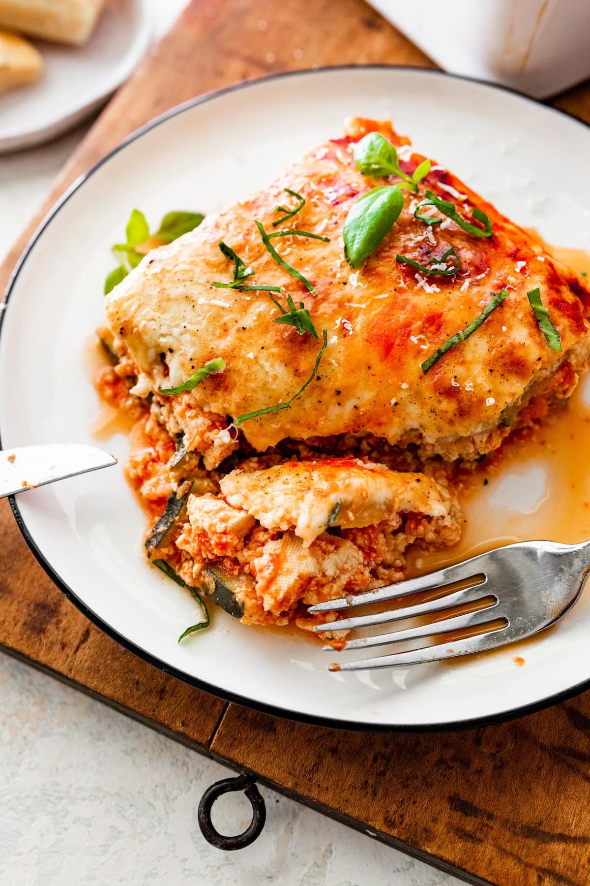 A serving of cheesy vegetarian tofu lasagna made with zucchini noodles on a white plate with a fork and a knife.