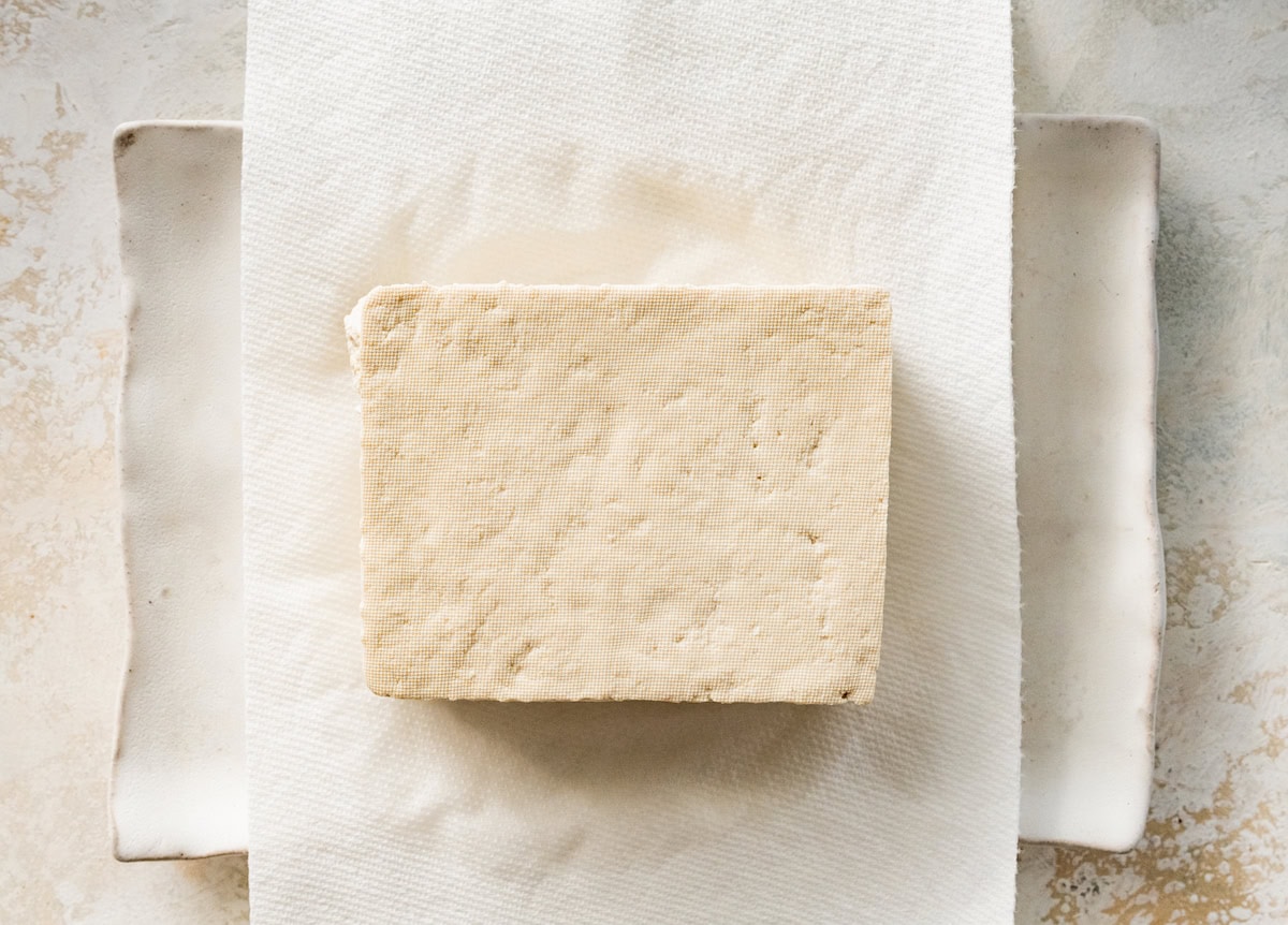 A block of tofu sits on a paper towel that is on a plate.