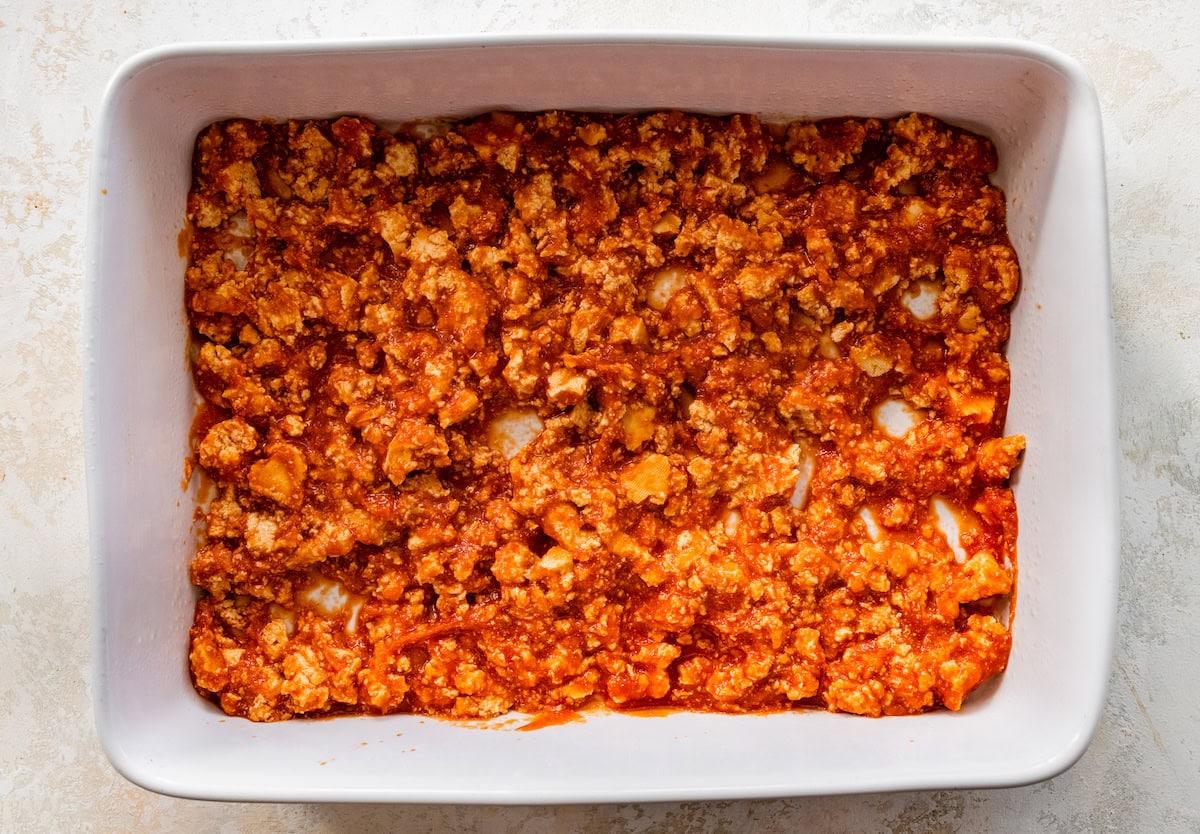 A layer of crushed tofu and marinara sauce in a square baking dish used to as the base of tofu zucchini lasagna.