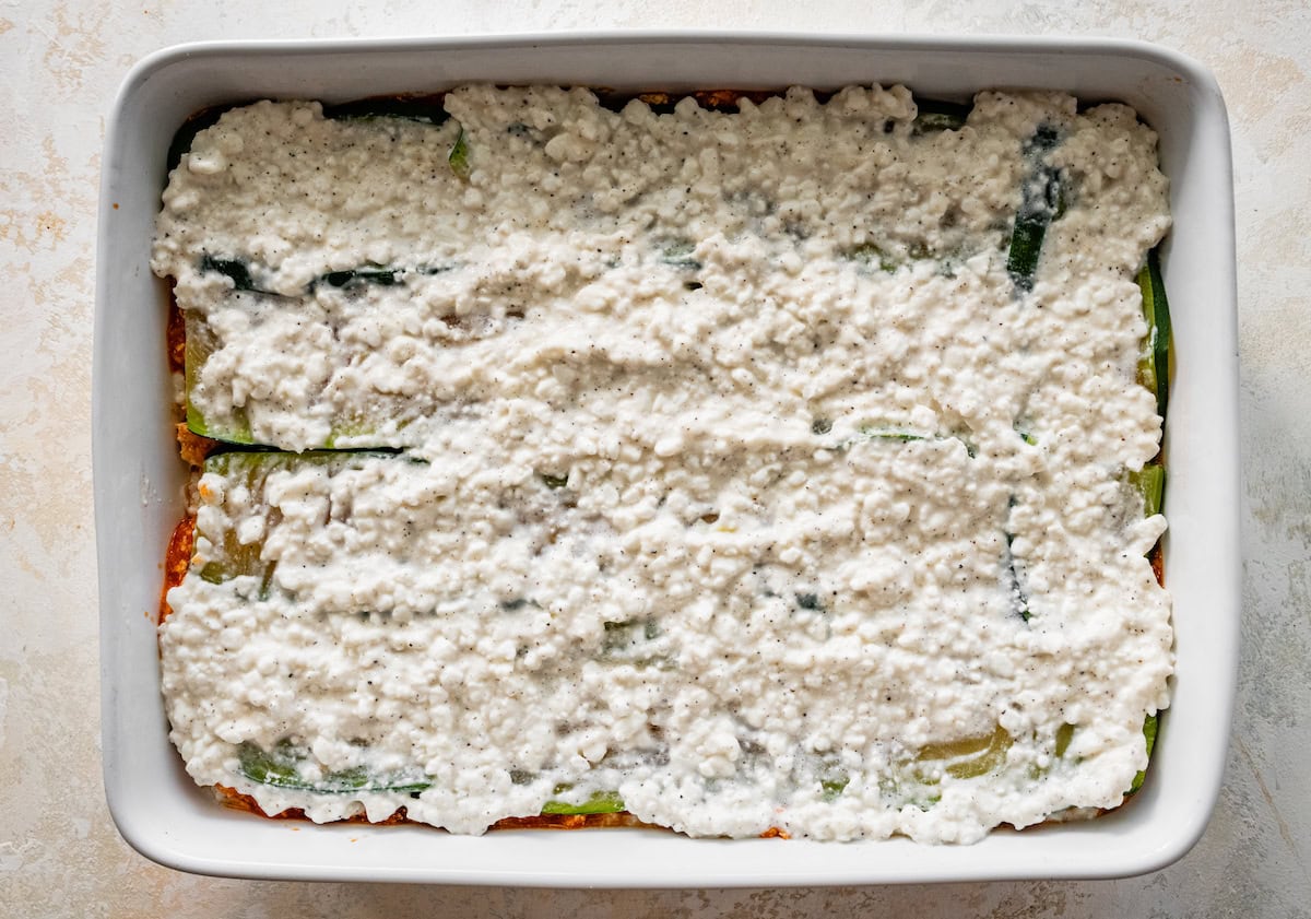 A layer of zucchini slices and cottage cheese on top of a marinara and tofu mixture in a square baking dish.