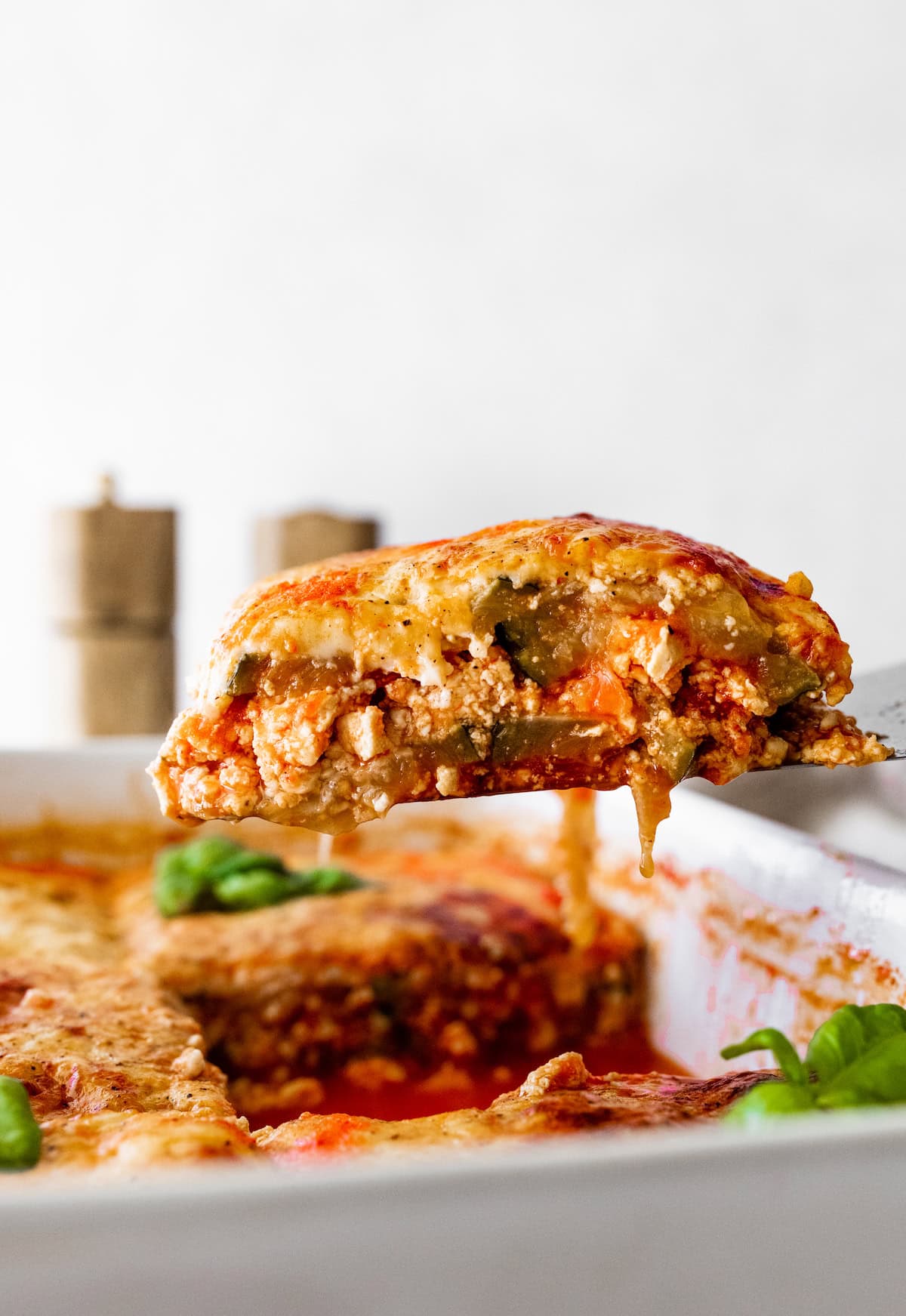 A serving of tofu zucchini lasagna being lifted from a baking dish by a metal spatula.