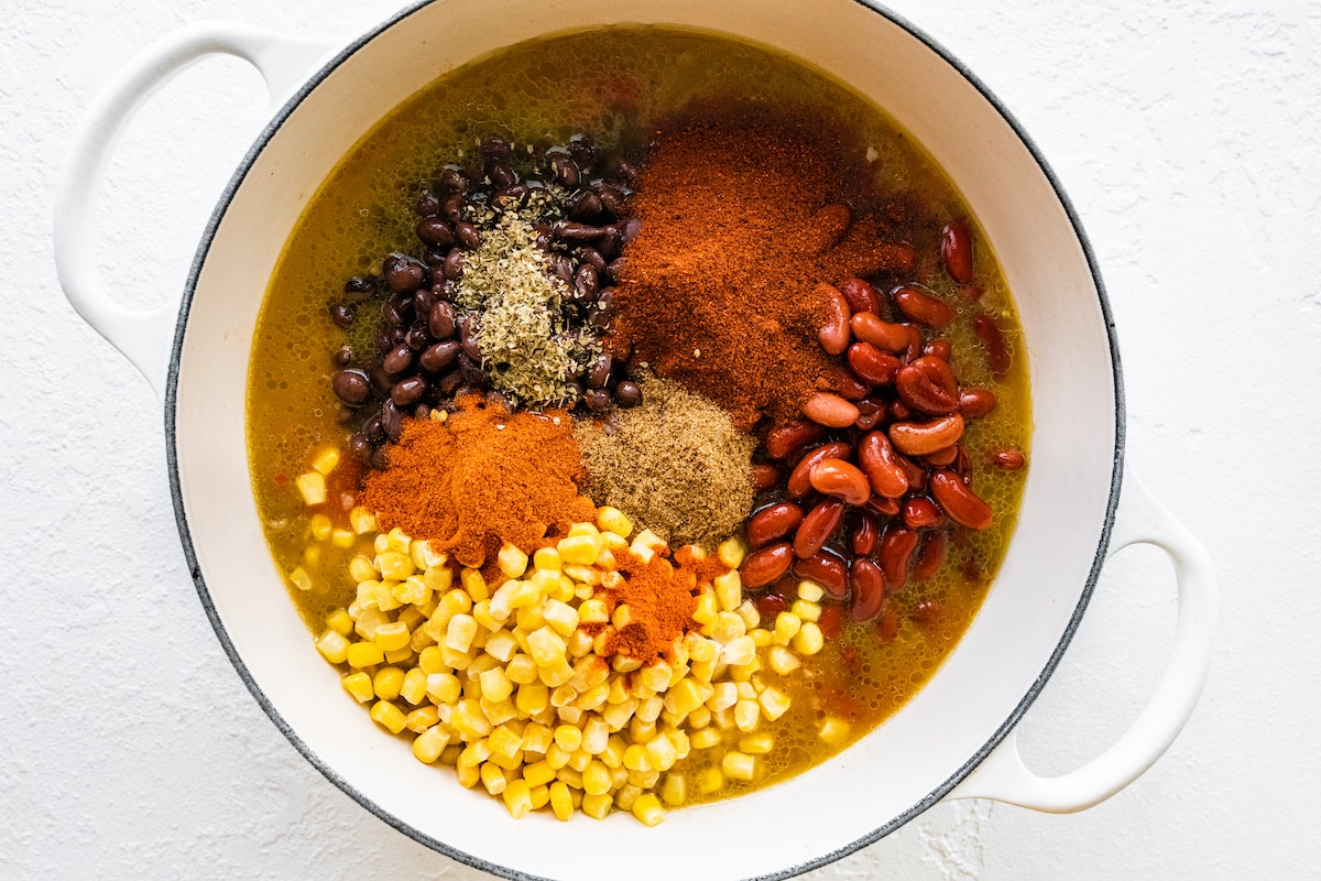 Turkey chili ingredients before being mixed together in a large white dutch oven. Ingredients include kidney beans, black beans, corn, broth, and plenty of spices.