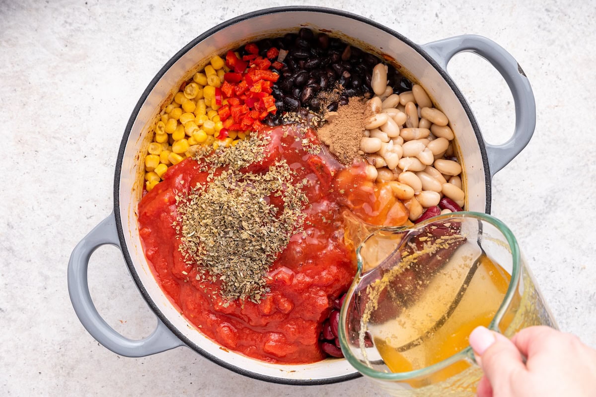 A womans hand pours vegetable broth into a large pot with multiple ingredients to make a vegetarian chili.