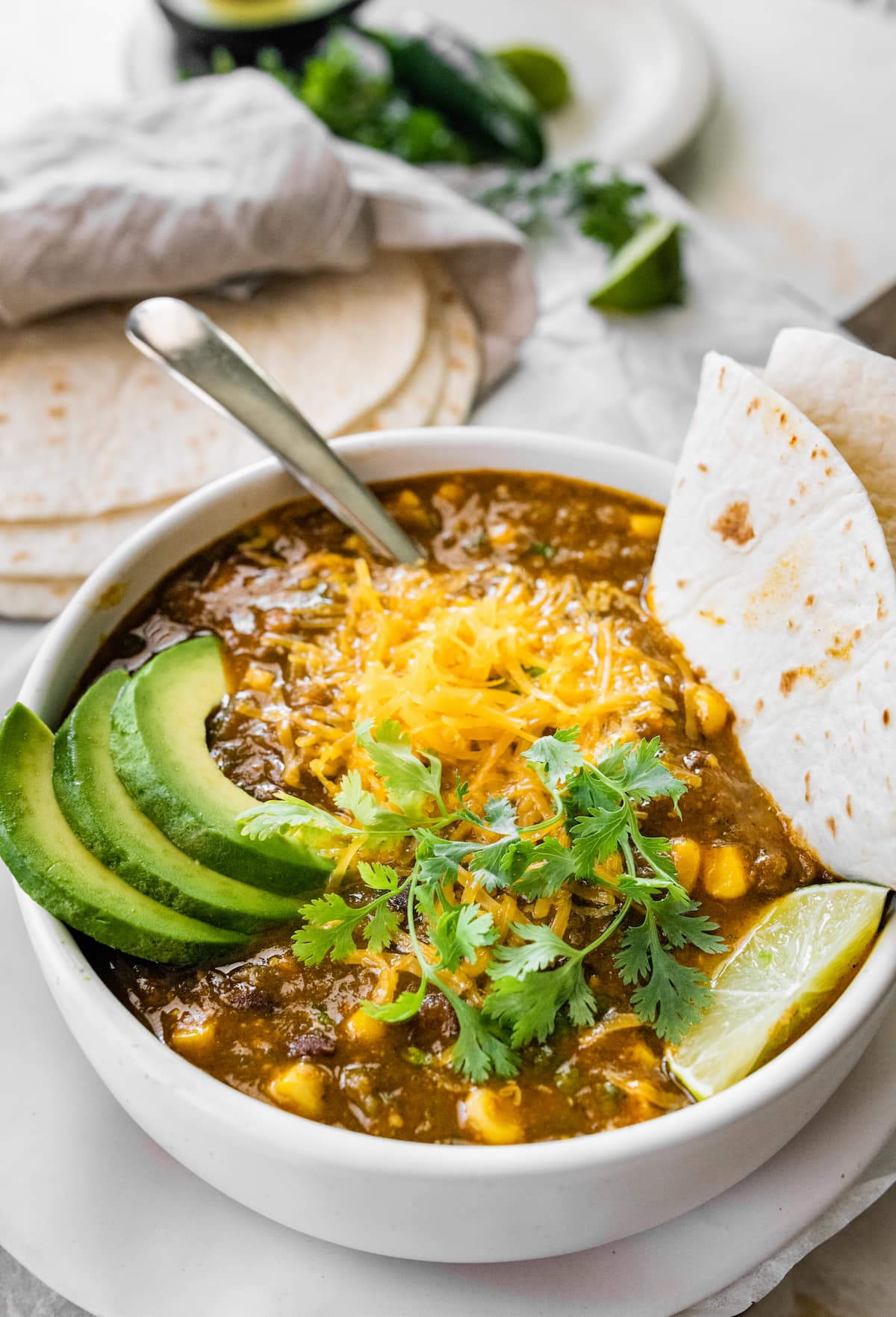 A bowl of vegetarian chili verde served with avocado, lime, cheese, cilantro and a tortilla.