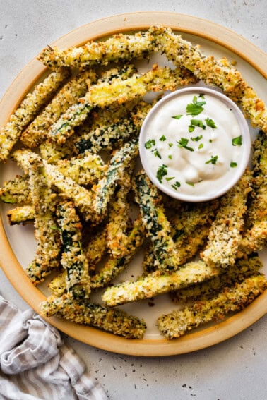 A large plate full of crispy zucchini fries covered in panko bread crumbs with a small dish of a dip.