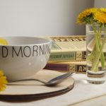 A white bowl with Good Morning text, dandelions in a glass, and books
