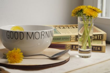 A white bowl with Good Morning text, dandelions in a glass, and books